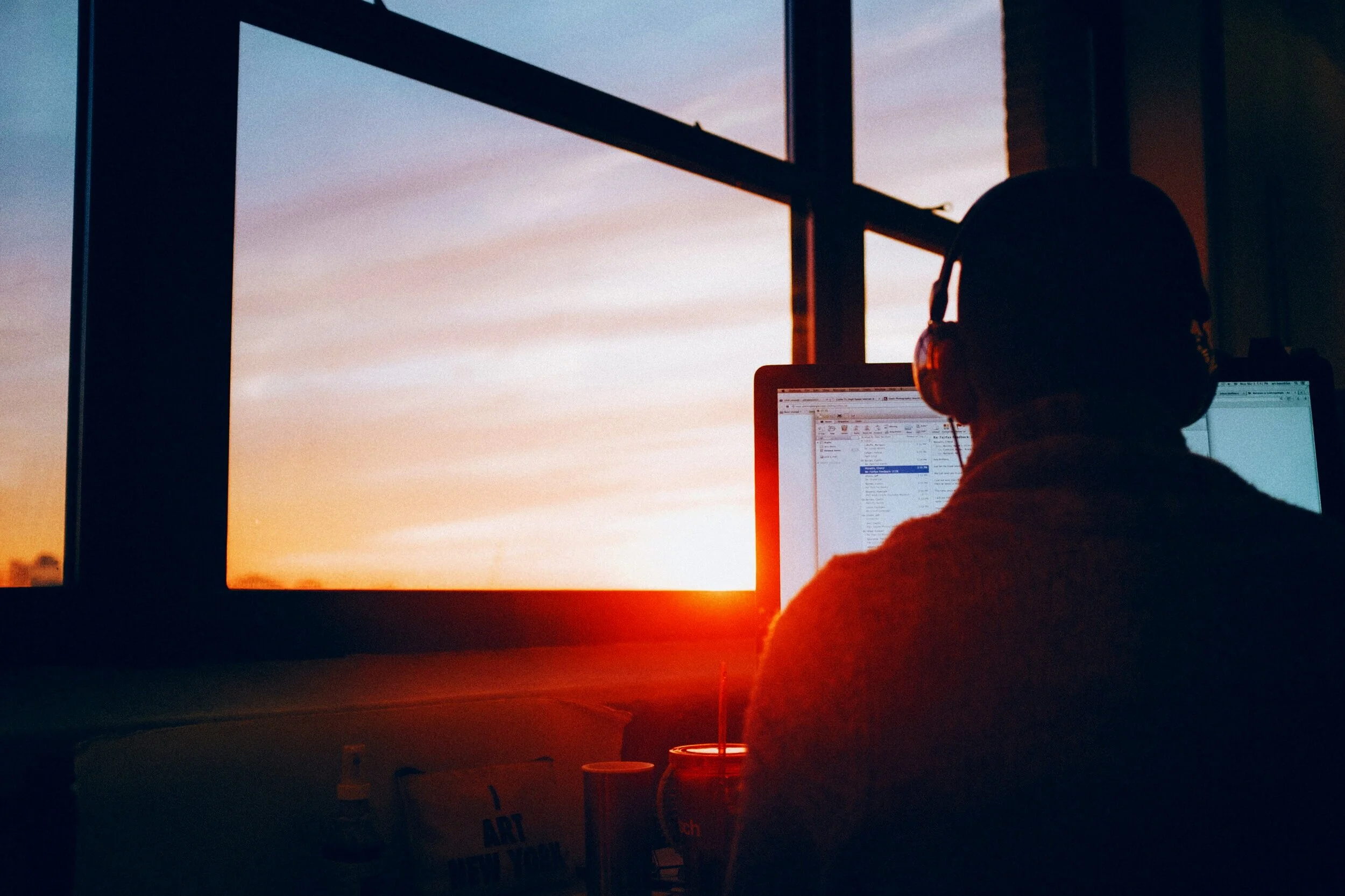 A person sitting at a desk wearing headphones, working on a computer with a sunset view through a large window in the background.