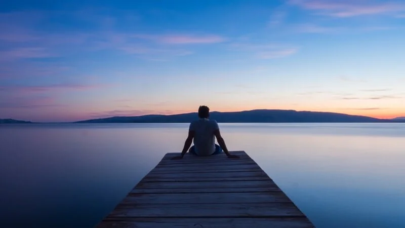A person sitting on a wooden dock by a calm body of water during sunset.