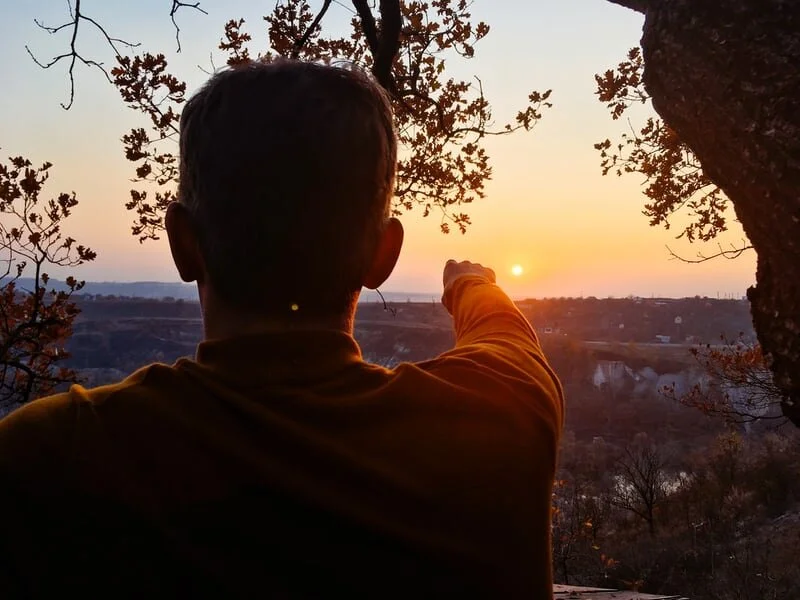 A person facing away, pointing towards the sunset in a natural outdoor setting with trees and hills.