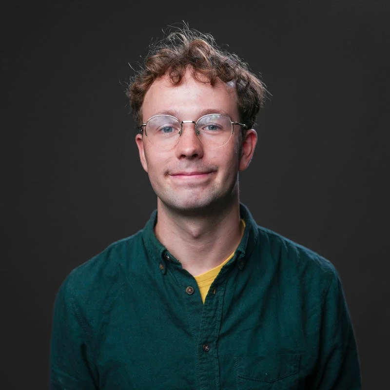 A young man with curly brown hair and glasses, smiling slightly, wearing a dark green button-up shirt over a yellow shirt, against a plain dark background.