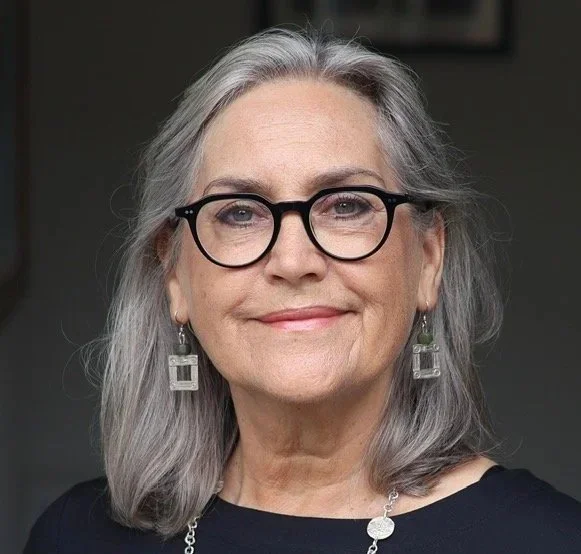 A portrait of a woman with shoulder-length gray hair, wearing black-rimmed glasses, earrings, and a black top, smiling slightly against a plain light background.