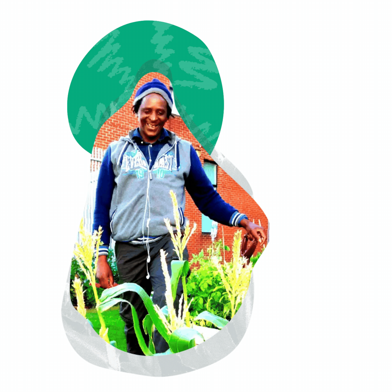 A smiling man wearing a gray jacket, blue hoodie, and beanie, standing in a garden with tall green plants in front of a brick house.