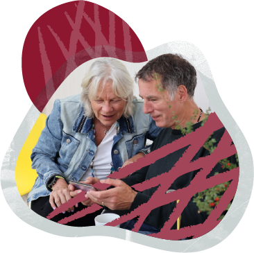 An elderly woman and a middle-aged man sitting outside, looking at a phone together, with a decorated Christmas tree in the background.