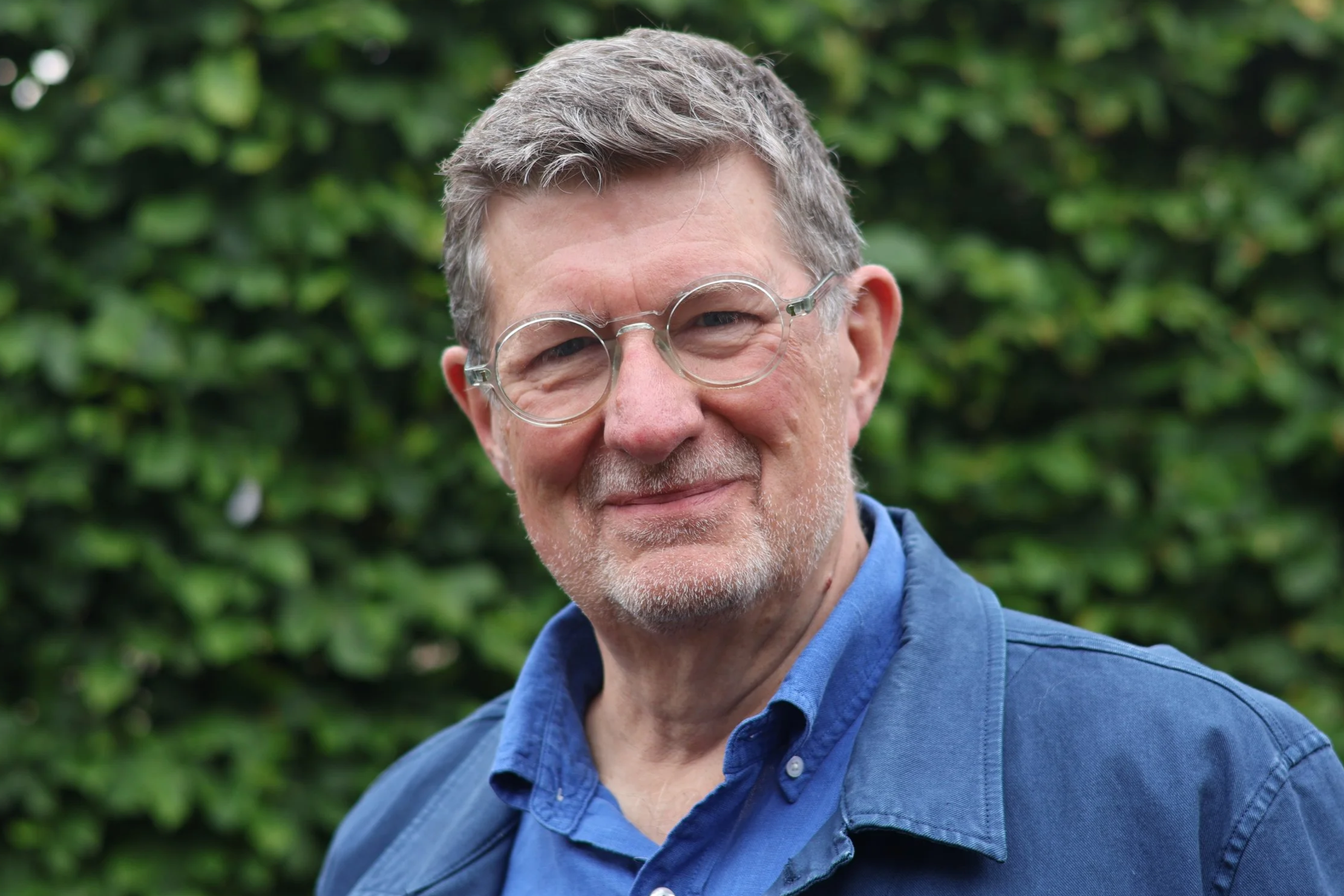 Portrait of an older man with gray hair, glasses, and a beard, wearing a dark turtleneck and a gray blazer, standing in front of a white wall with vertical stripes.