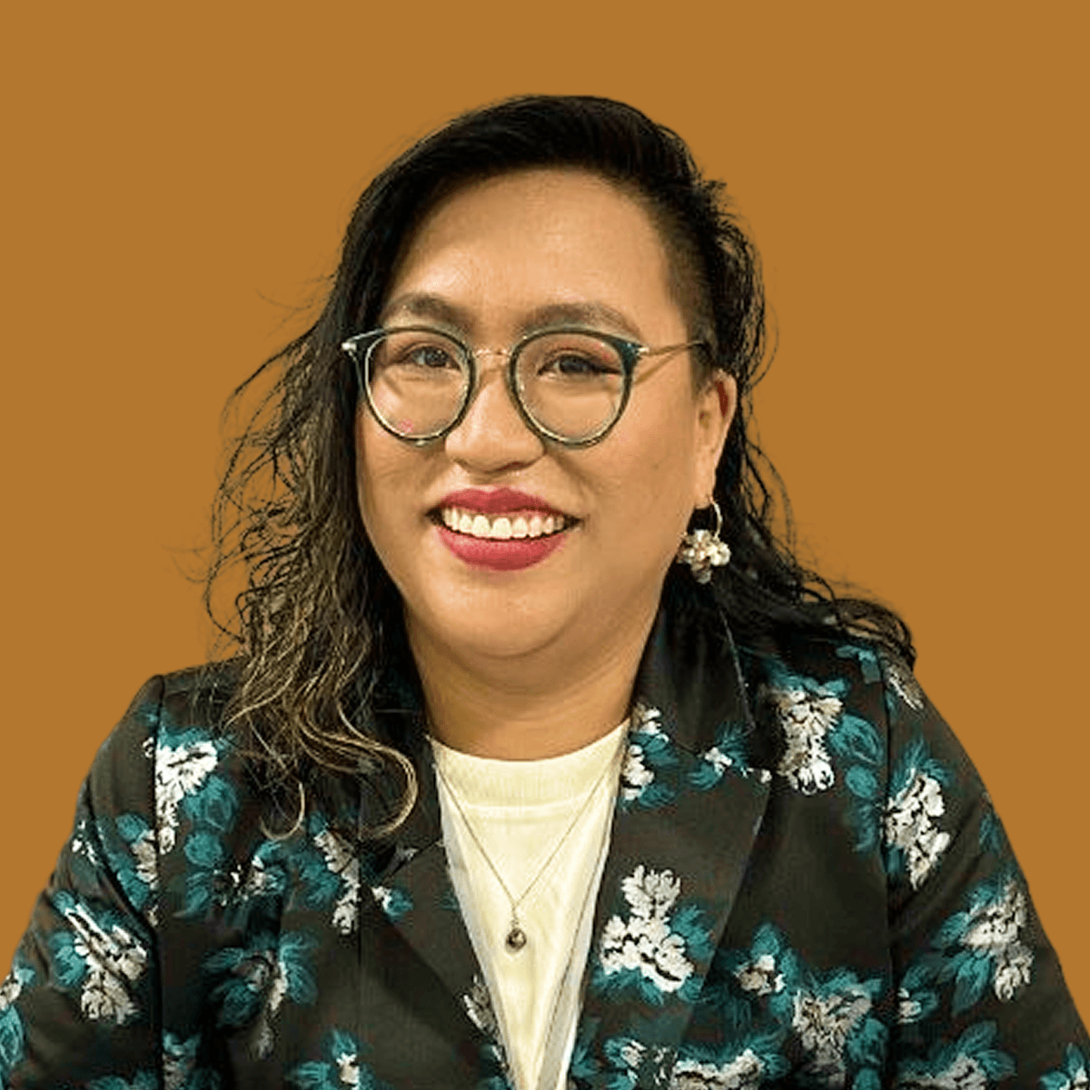 A woman with glasses and earrings, smiling, wearing a floral blazer and a cream-colored top against a brown background.