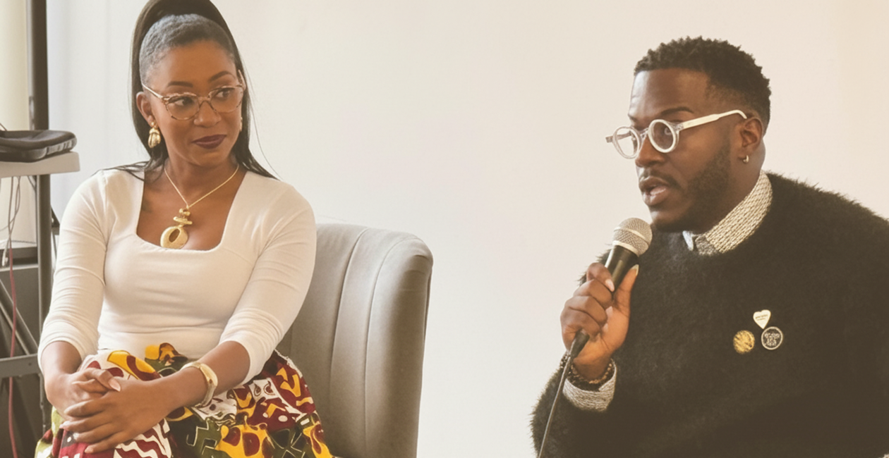 A woman and a man are seated indoors, having a conversation. The woman is wearing glasses, a white top, and colorful patterned skirt, while the man is speaking into a microphone, dressed in a black sweater with pins, and wearing white-framed glasses.