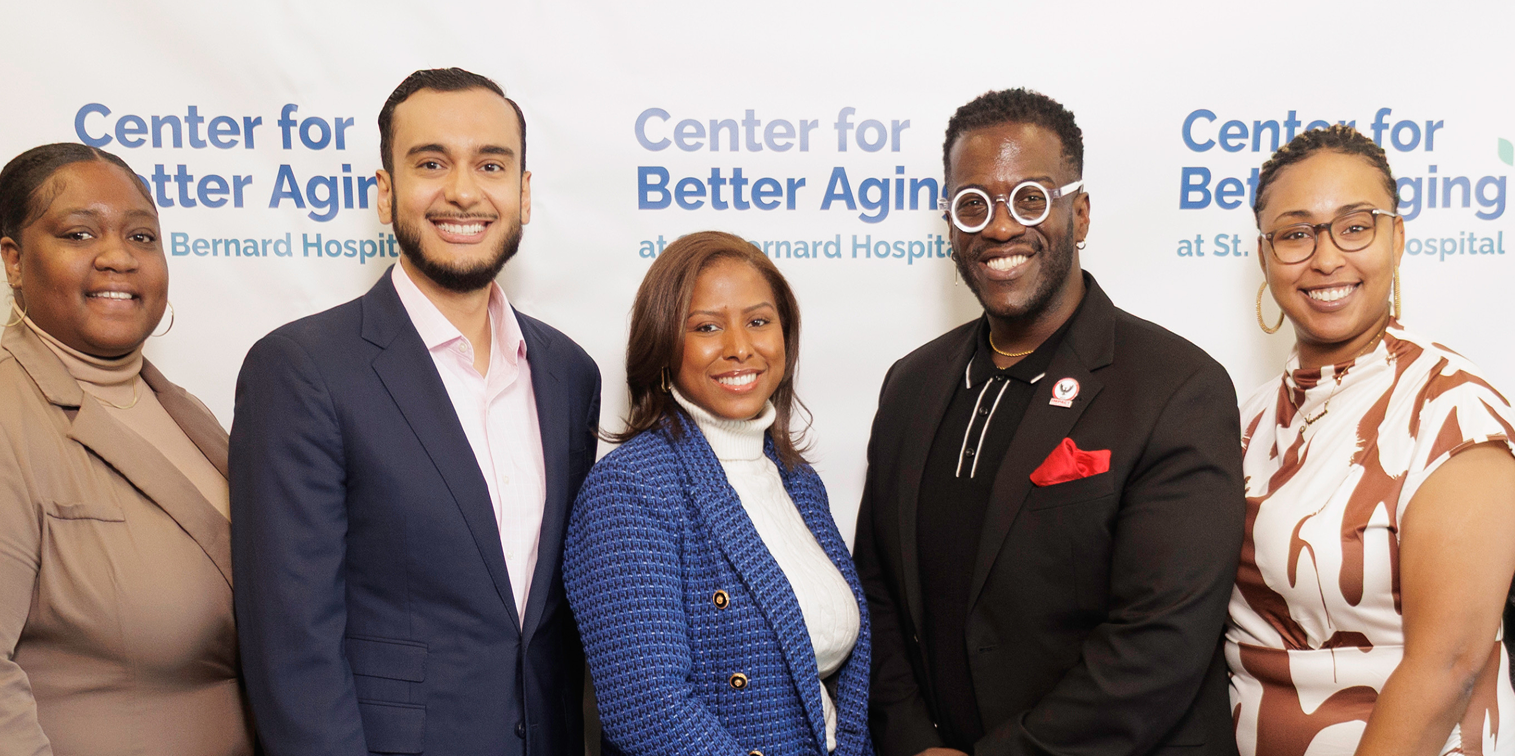 Group of five diverse professionals standing and smiling in front of a banner that reads 'Center for Better Aging at St. Bernard Hospital'.