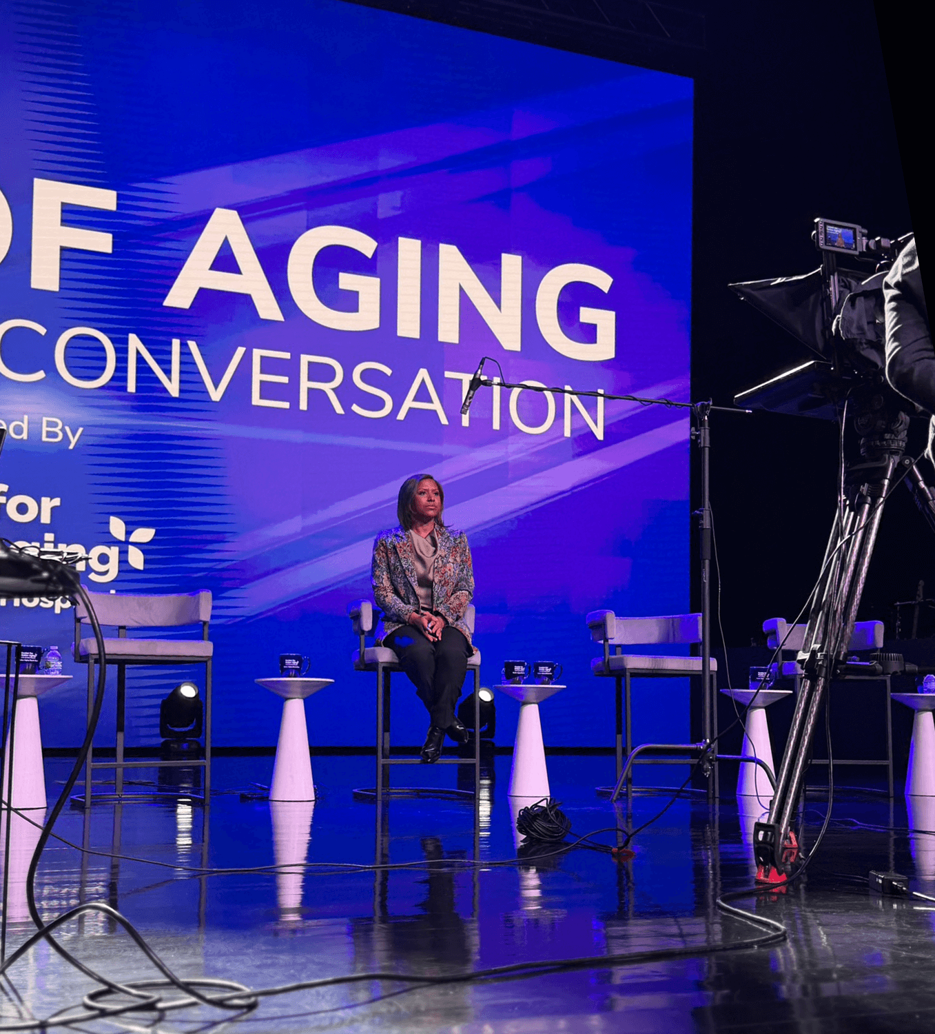 A woman sitting on a stage during a panel discussion at an event titled 'Aging Conversation' with professional camera equipment and lighting set up around her.