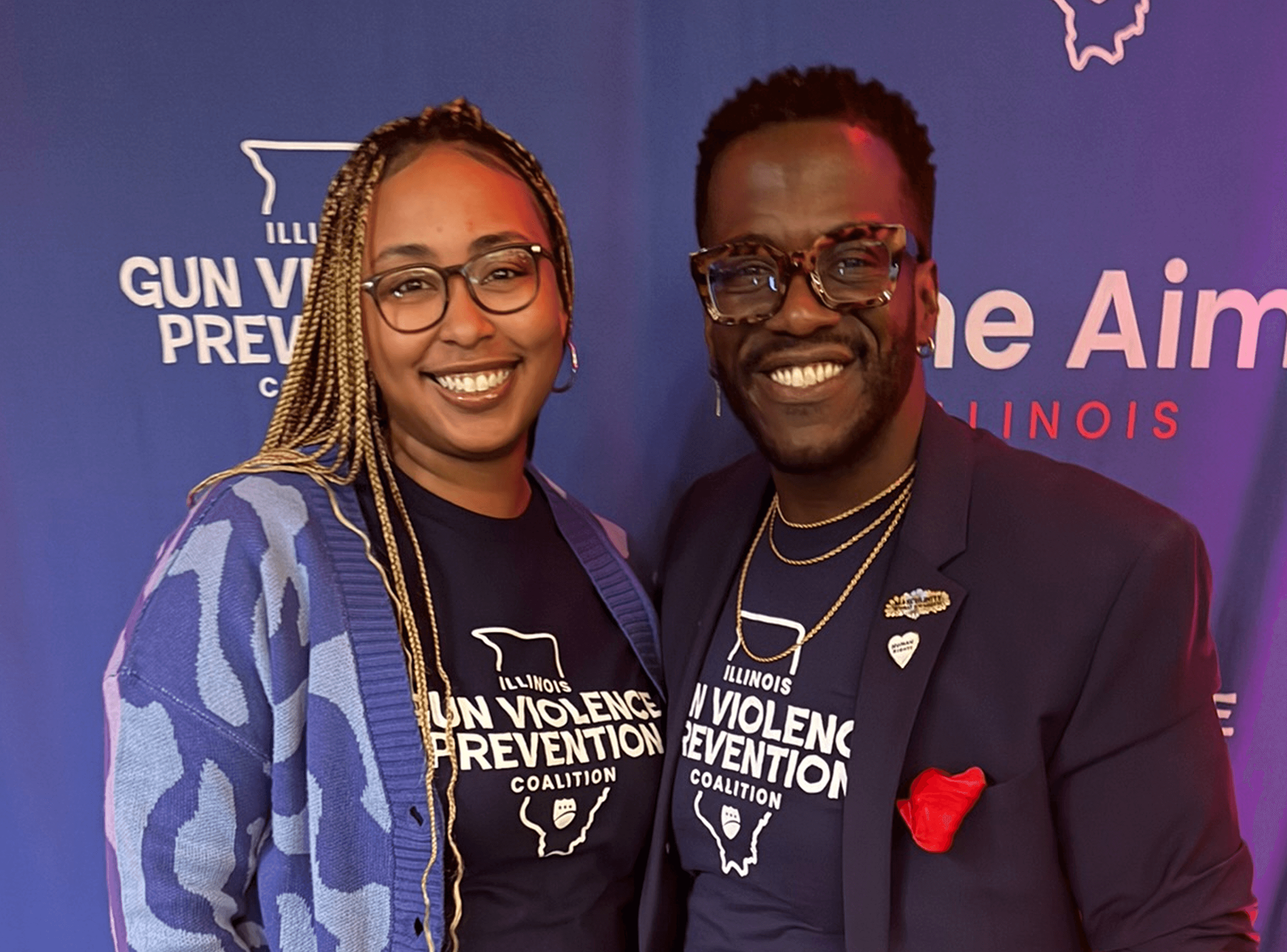 Two smiling individuals wearing matching Illinois violence prevention coalition shirts at a community event, standing in front of a blue backdrop with event text.