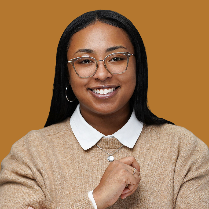 A young woman with black hair, glasses, and hoop earrings, smiling, wearing a beige sweater over a white collared shirt, with a necklace, posed against a solid brown background.