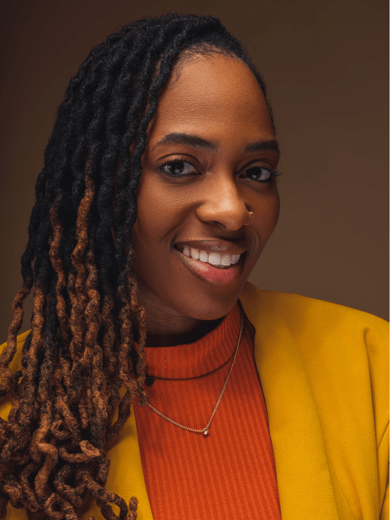 A woman with dark skin and long dreadlocks smiling, wearing a yellow blazer, a red ribbed top, a silver necklace, and a nose ring.