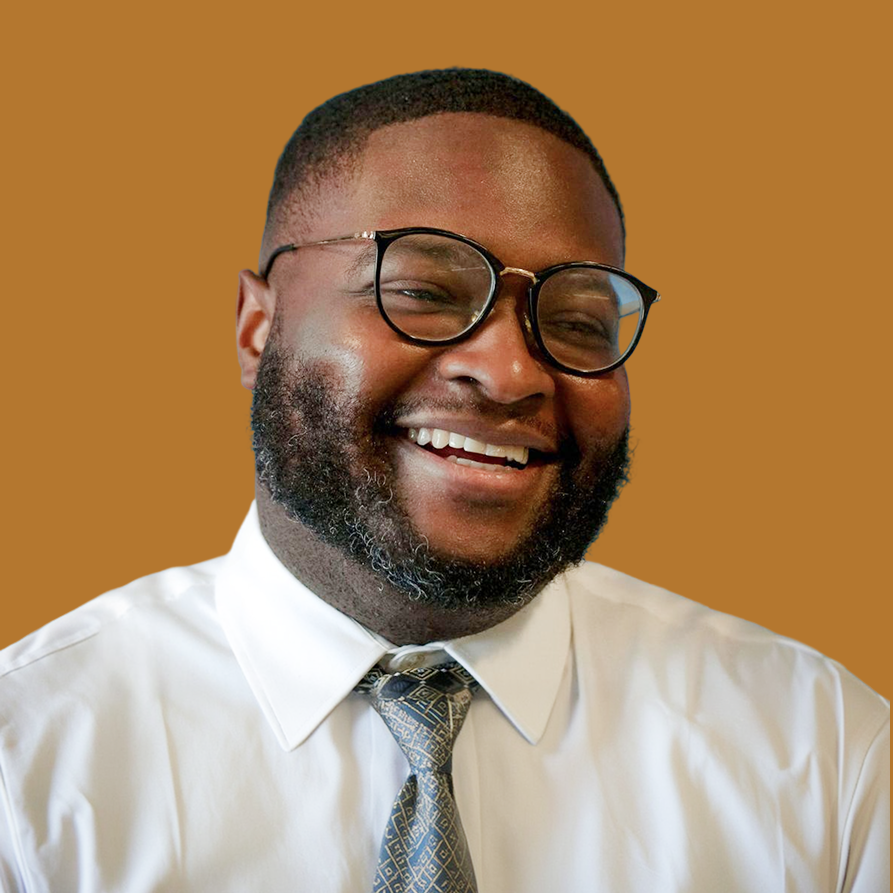 Portrait of a smiling man with glasses, wearing a white shirt and patterned tie, against a plain brown background.