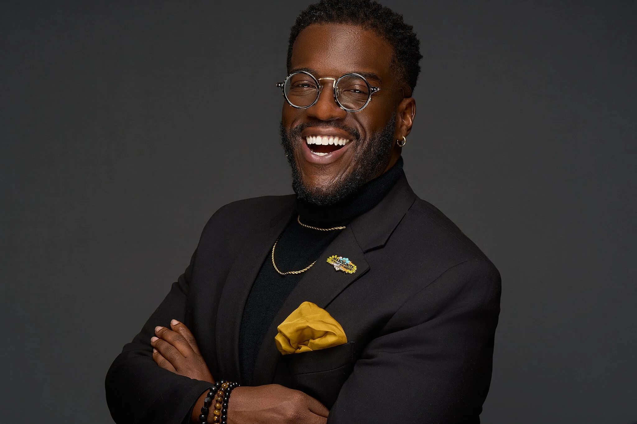 A smiling man with glasses, a beard, and gold jewelry, wearing a black blazer with a yellow pocket square, standing against a dark background.
