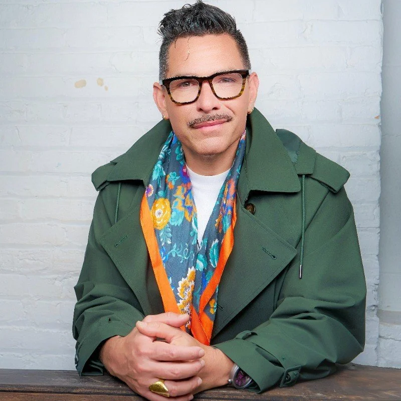 A man with short curly hair, wearing glasses, a green trench coat, and a colorful patterned scarf, sitting at a wooden table with a white brick wall in the background.