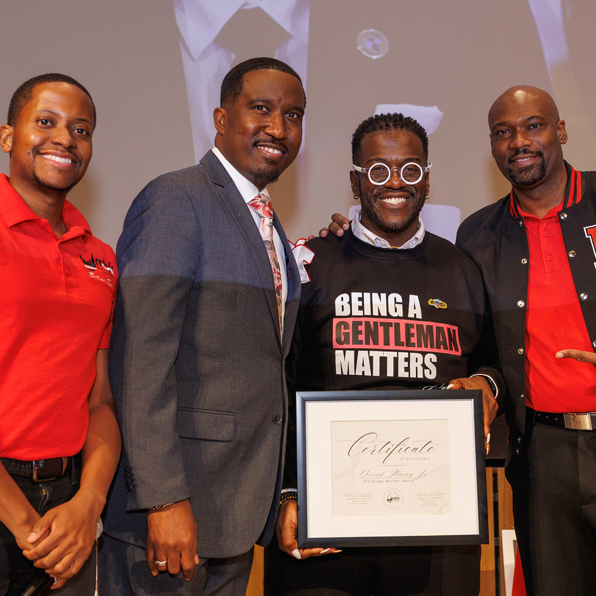 Four men standing together at an award ceremony, with the man second from the right holding a framed certificate. The man second from the right is wearing glasses and a black shirt with the text 'BEING A GENTLEMAN MATTERS.' The man on the far left is wearing a red polo shirt, and the man on the far right is dressed in a black and red jacket. The background shows a screen with some graphics.