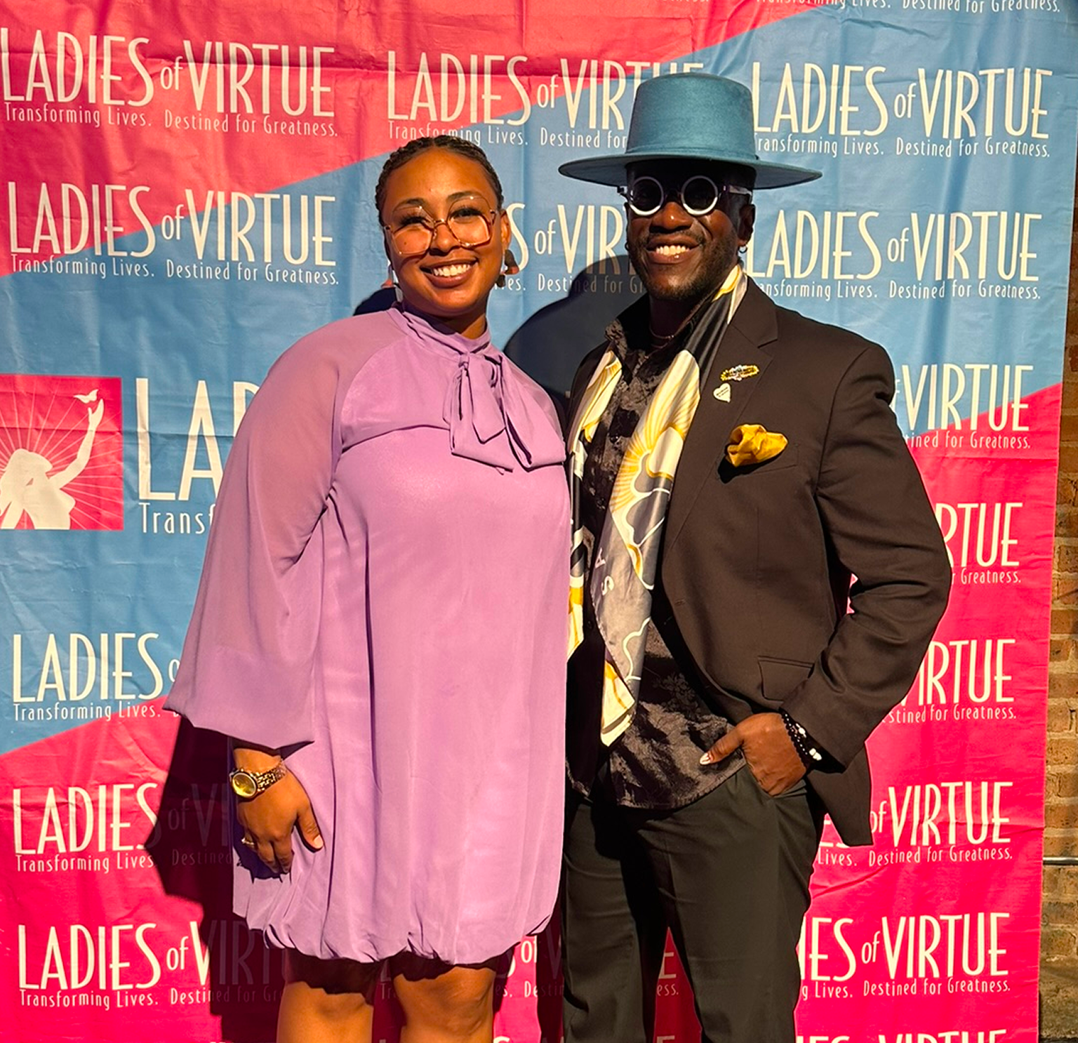 A woman and a man posing together at a Ladies of Virtue event. The woman is wearing a lavender dress with a bow at the neck, glasses, and a gold watch. The man is wearing a dark suit with a colorful tie, a blue hat, glasses, and has a yellow pocket square. They are standing in front of a red and blue Ladies of Virtue backdrop.