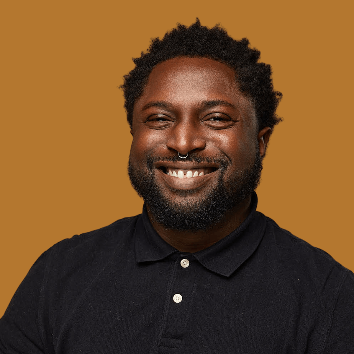 Portrait of a smiling Black man with a beard and septum piercing, wearing a black polo shirt, against a brown background.