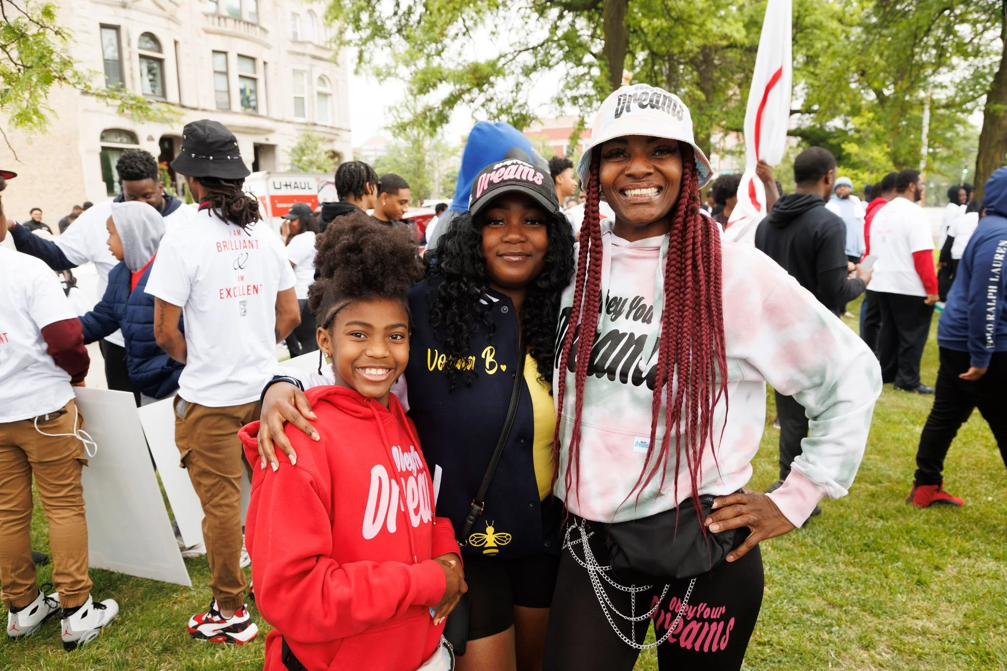 Three women smiling and posing for a photo at an outdoor event, with a crowd in the background and trees surrounding the area.