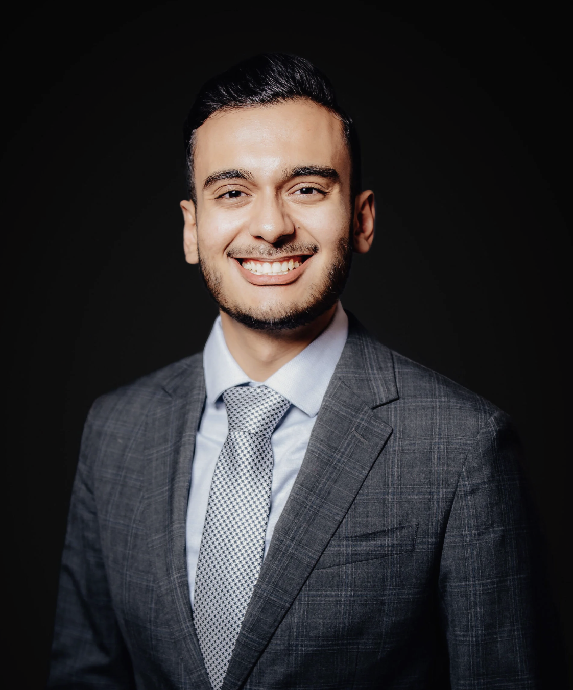 Professional headshot of a smiling man in a gray plaid suit with a light blue shirt and patterned tie, against a dark background.