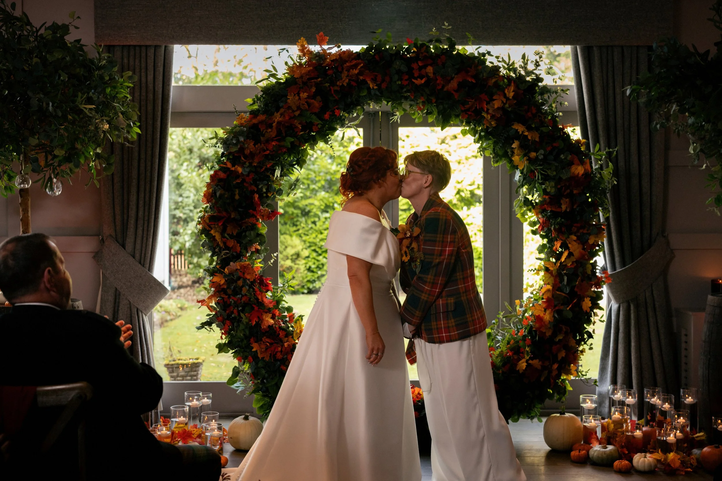 two brides kiss at the alter on their autumnal themed wedding in front of a large round archway covered in autumnal florals and surrounded by candles and pumpkins.
