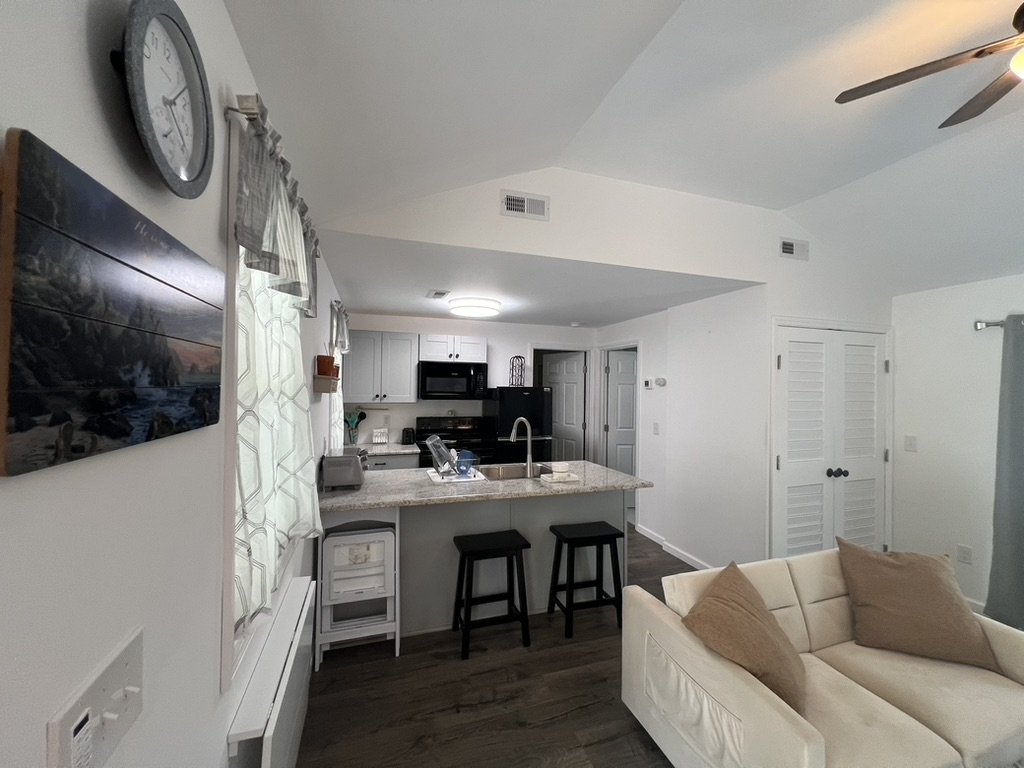 Living room and kitchen area with white walls and dark wood flooring. The living room has a white sofa with brown pillows. The kitchen has a granite counter with two black stools, white cabinets, black appliances, and a microwave. There is a clock on the wall and some curtains on the window.