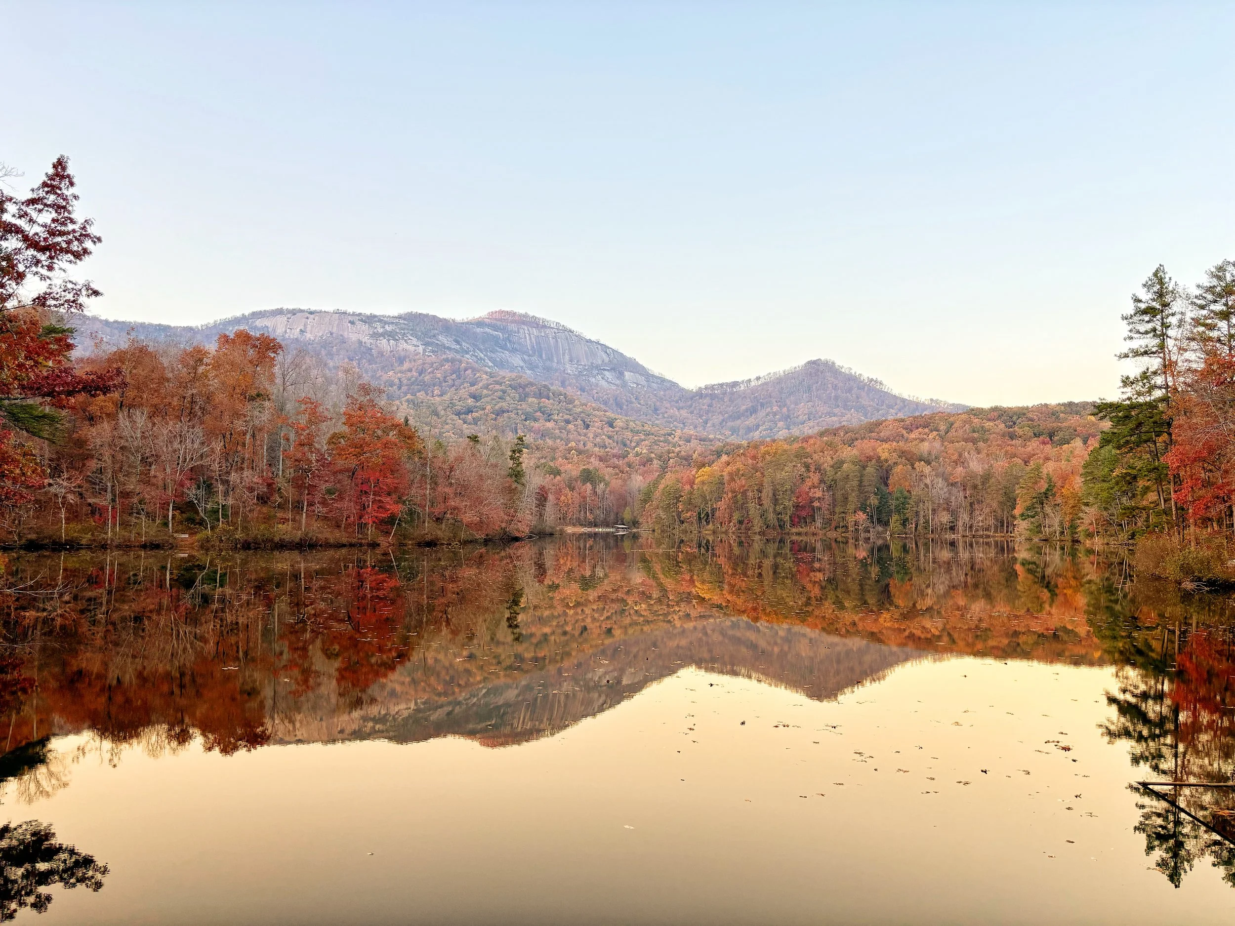 A peaceful lake surrounded by autumn-colored trees and mountains in the background, with a clear sky.