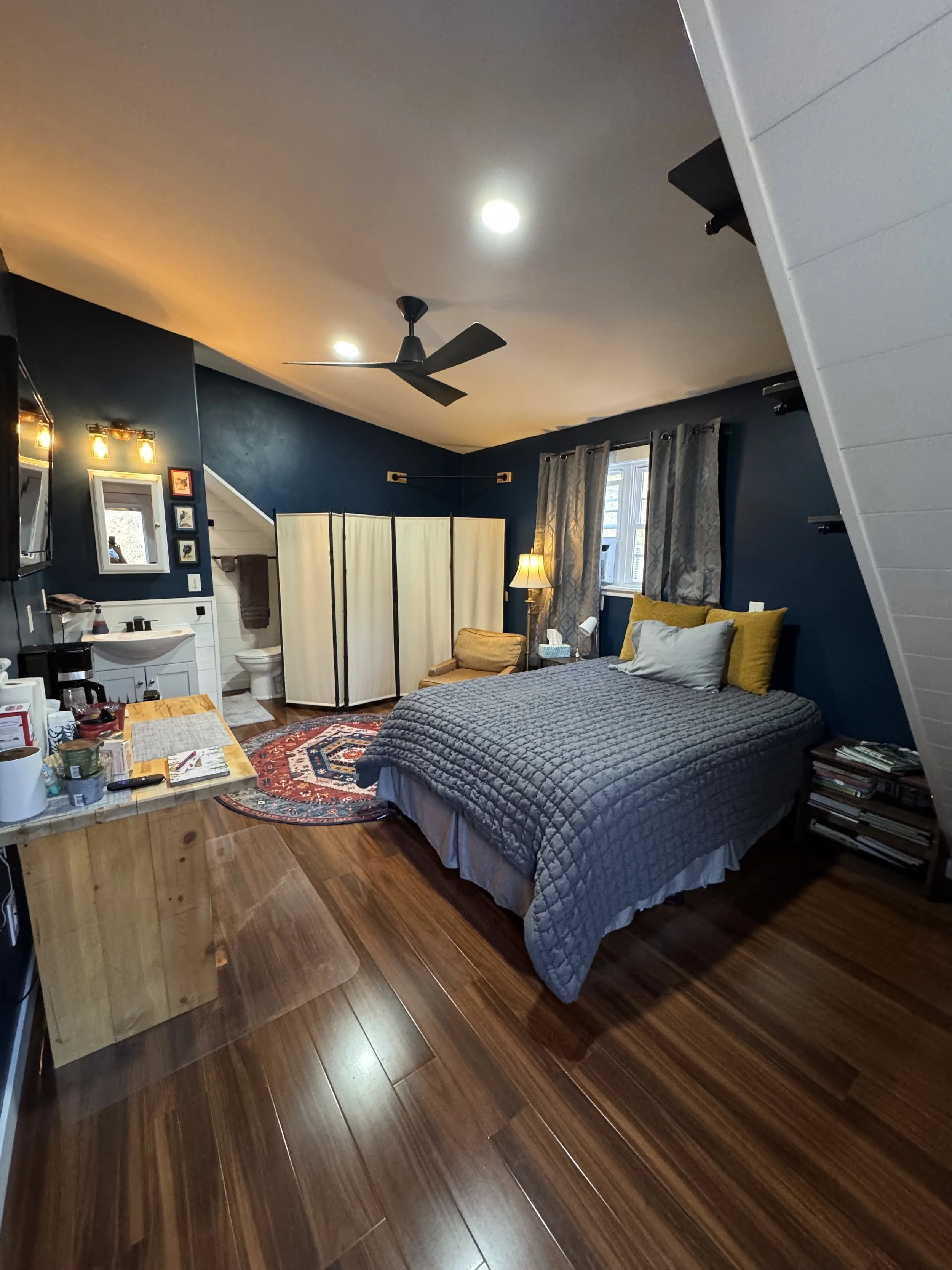 A cozy bedroom with dark blue walls, a wooden floor, a bed with gray quilt and pillows, a nightstand with a lamp, a window with curtains, a small bathroom area with a shower screen, a chair, and a folding room divider.