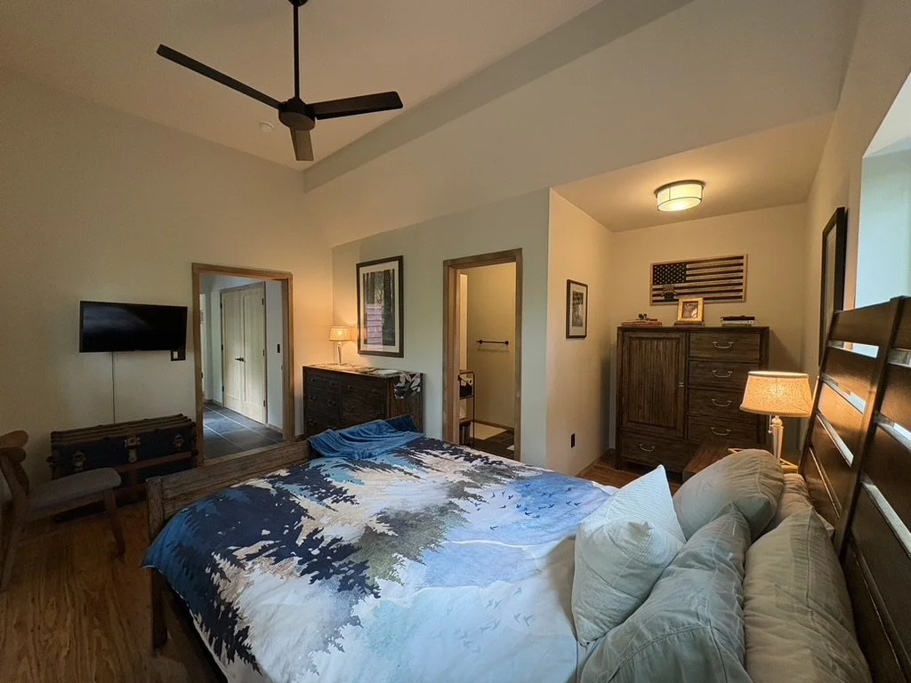 A bedroom with a ceiling fan, a mounted television, a bed with a nature-themed quilt, wooden furniture, and a wall decorated with an American flag.