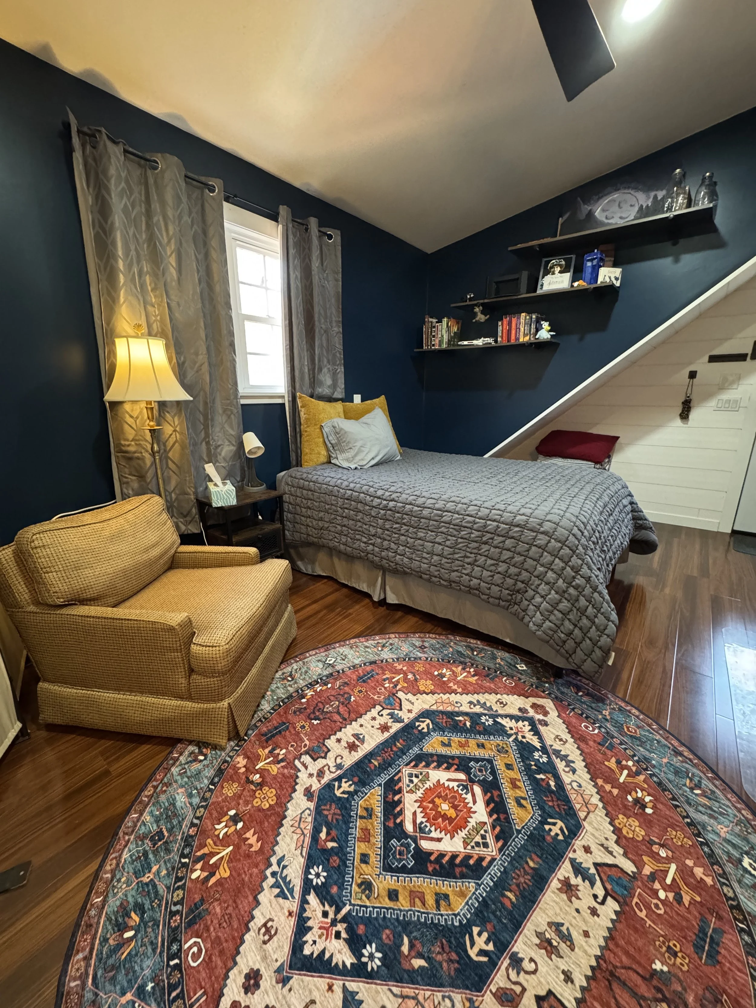 A cozy bedroom with dark blue walls and a sloped ceiling. It features a bed with a gray quilt and yellow pillows, a wooden side table with a lamp, a small brown armchair, and a vibrant patterned rug on wooden floor. Two shelves with books and decorative items are mounted on the wall above the bed, alongside a window with gray curtains.