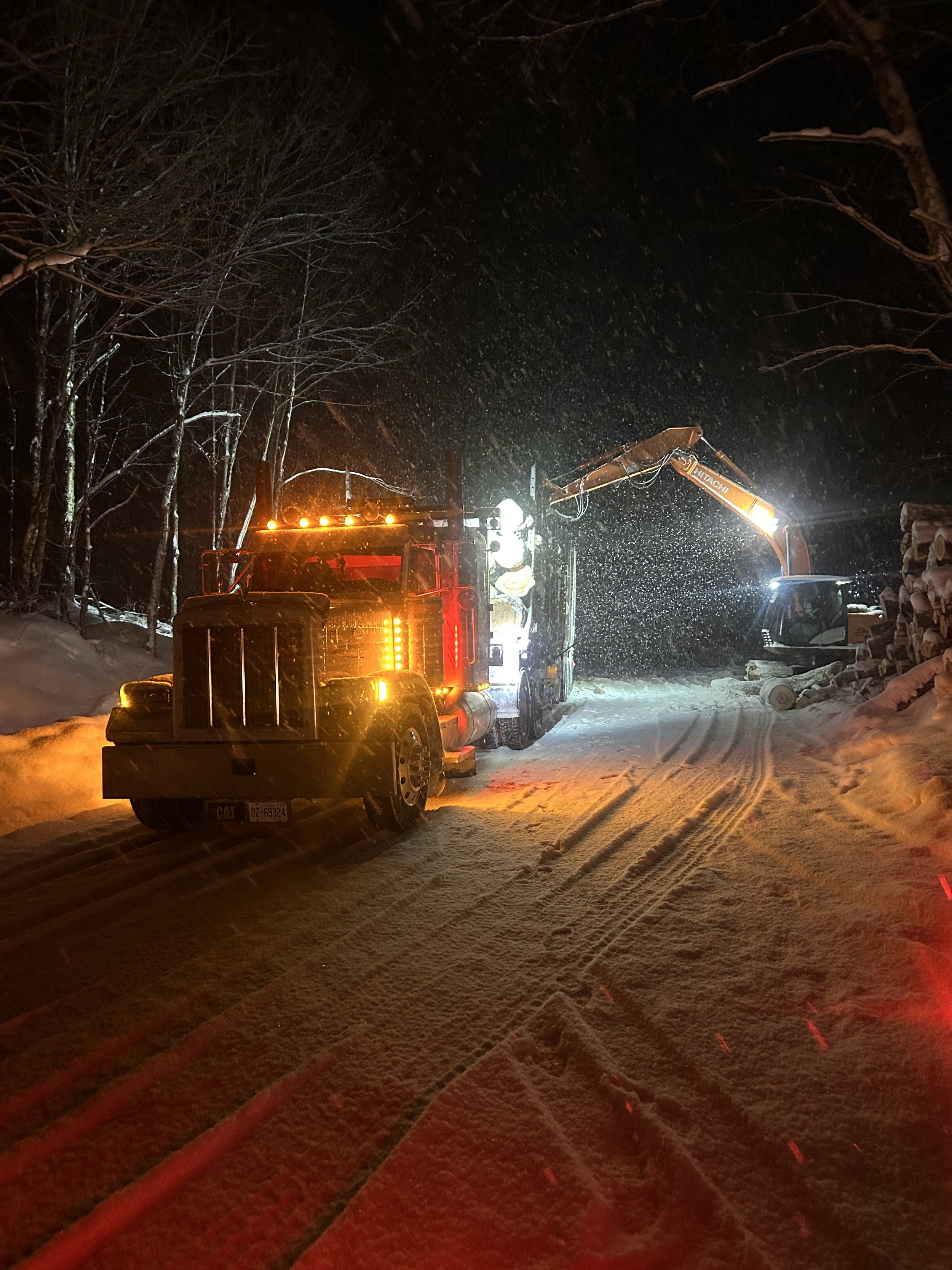 A snow-covered road at night with a work truck and a crane, illuminated by the truck's lights and snowfall.