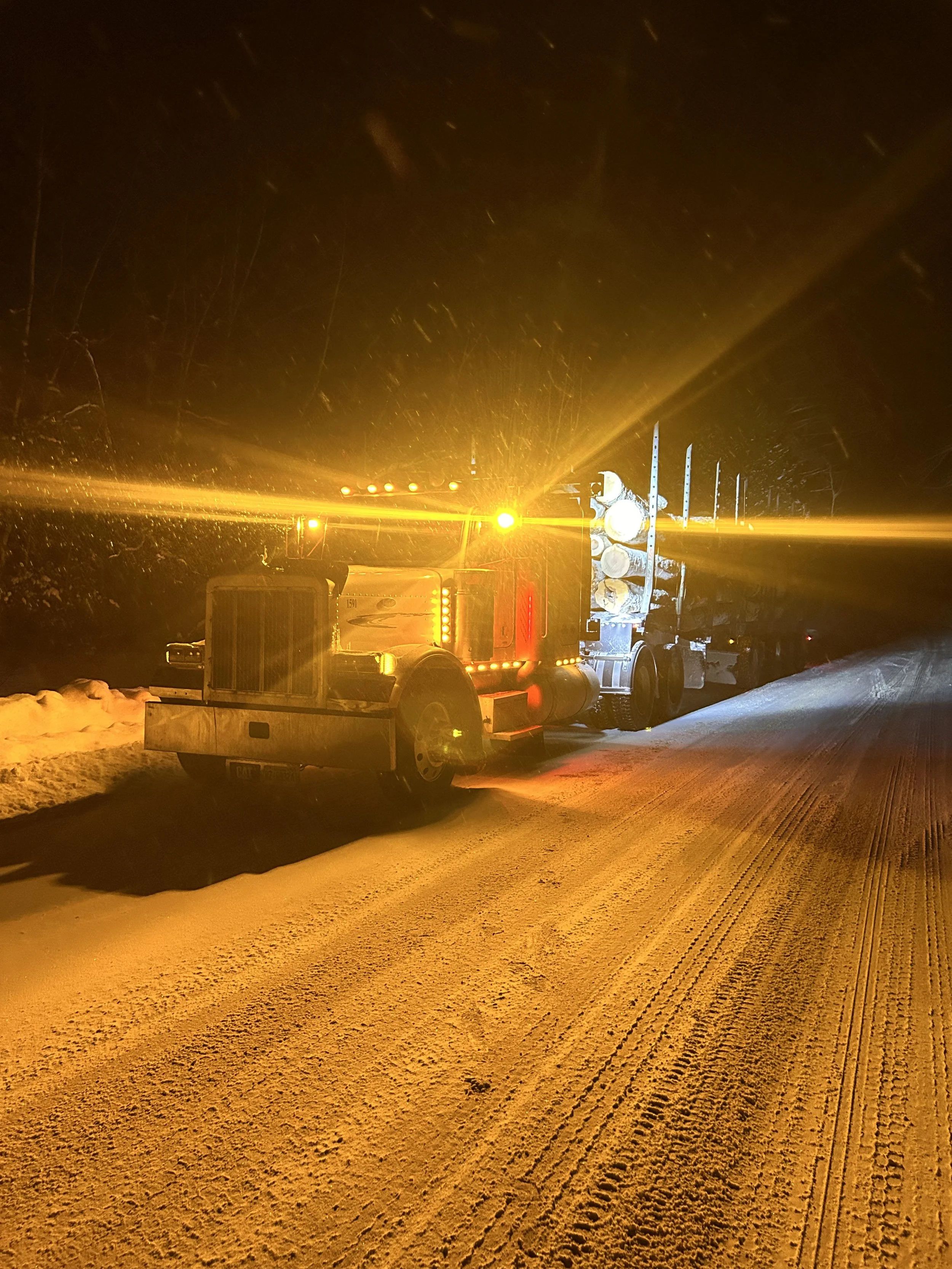 A brightly lit yellow snow plow truck working on a snowy road at night, with falling snow visible.
