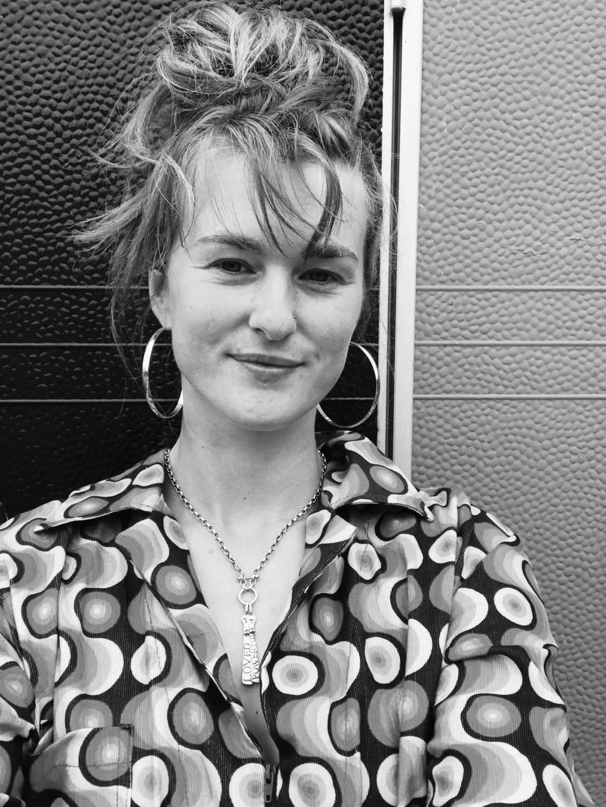 Black and white photo of a woman with tousled hair wearing large hoop earrings, a vintage patterned shirt and a charm necklace, standing in front of textured walls.