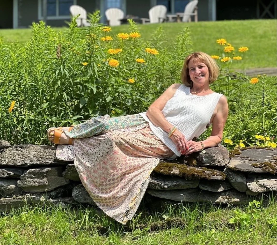 Carla Cross lying on a stone wall in a garden with yellow flowers, smiling at the camera.