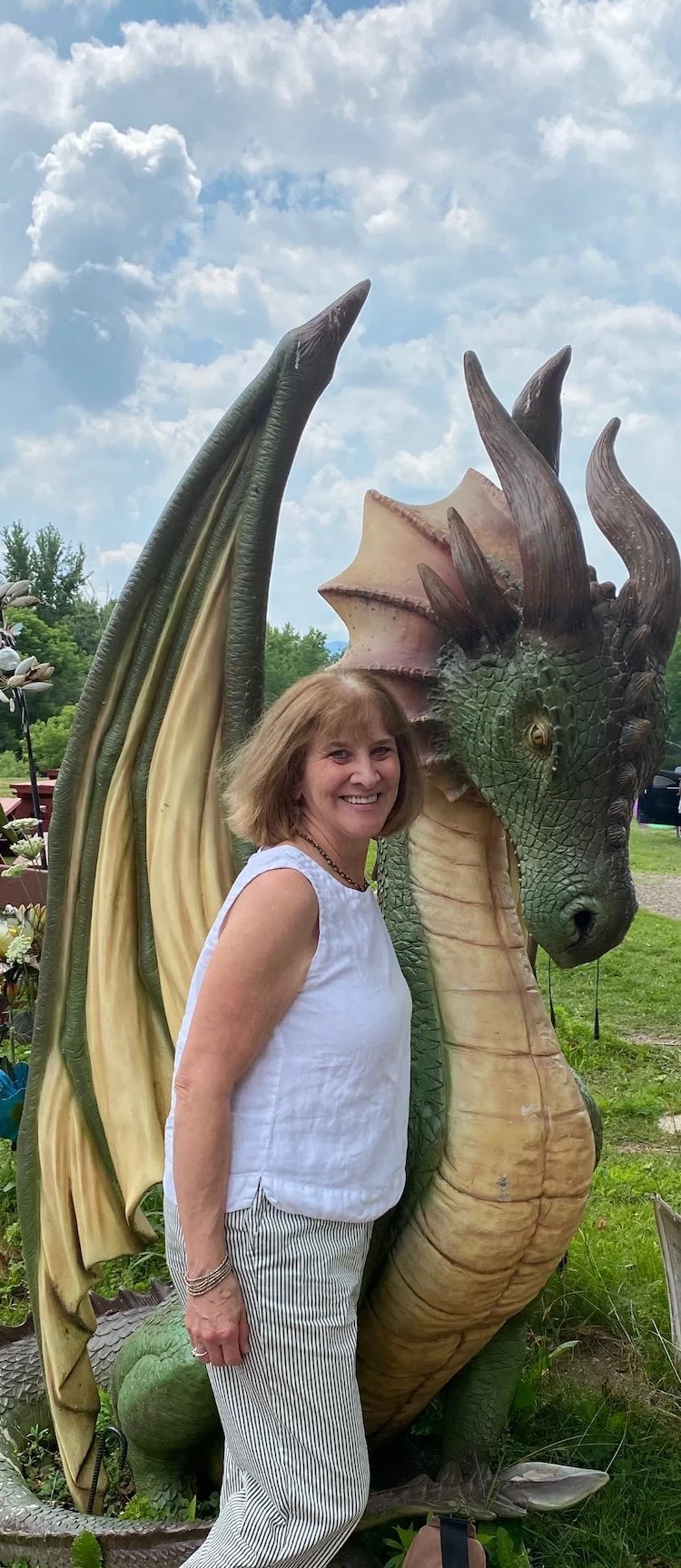 Carla Cross smiling and standing next to a large, colorful dragon sculpture outdoors on a cloudy day.