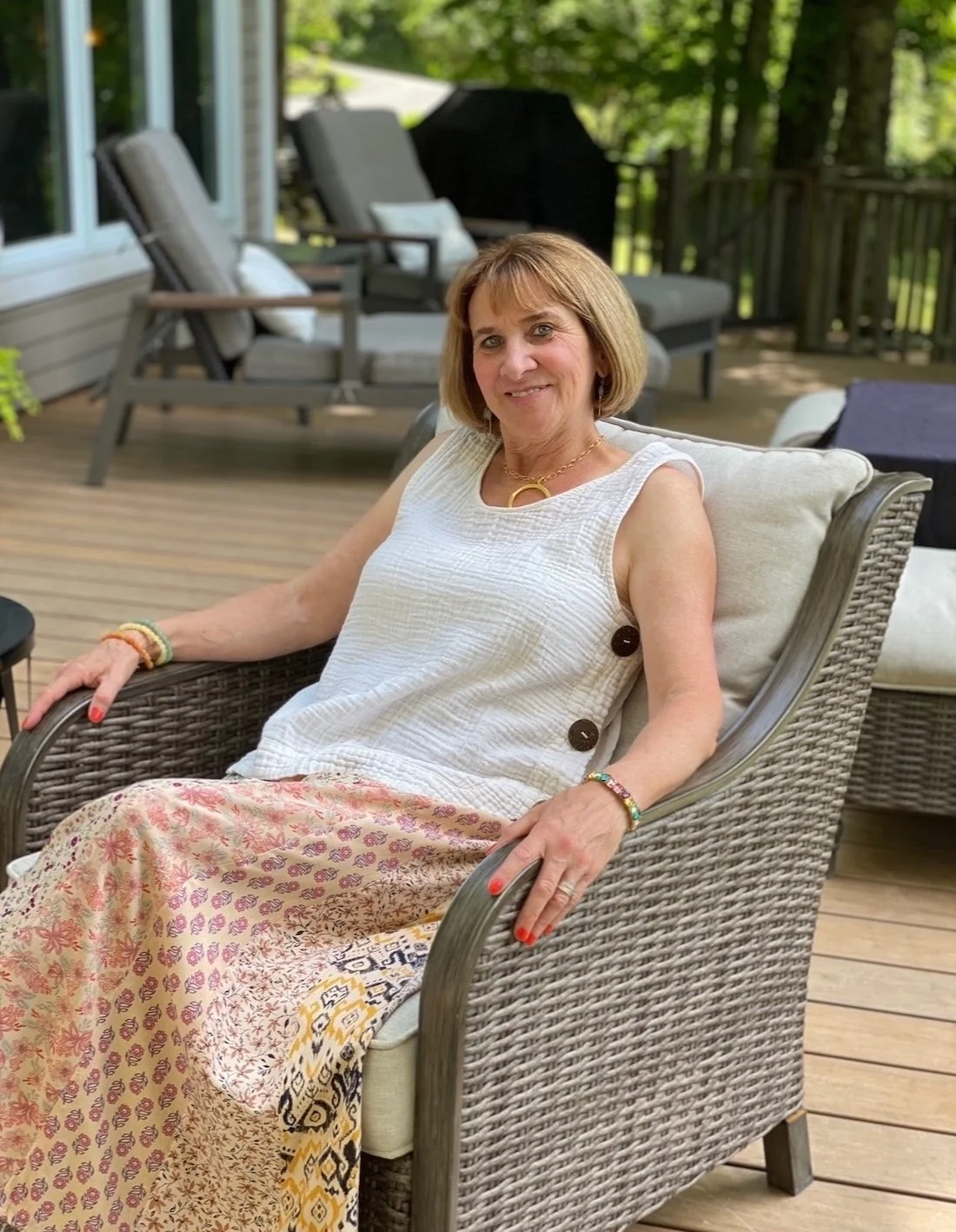 Carla Cross, owner of Passion and Purpose, sitting on a wicker chair on a wooden deck with outdoor furniture and trees in the background.