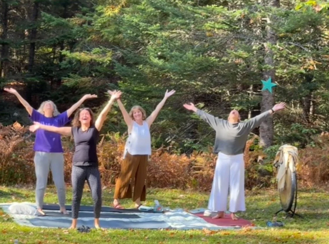 Four women and one man standing outdoors on a blanket in a forest, with their arms raised and eyes closed, appearing joyful and meditative, with trees and autumn foliage in the background.