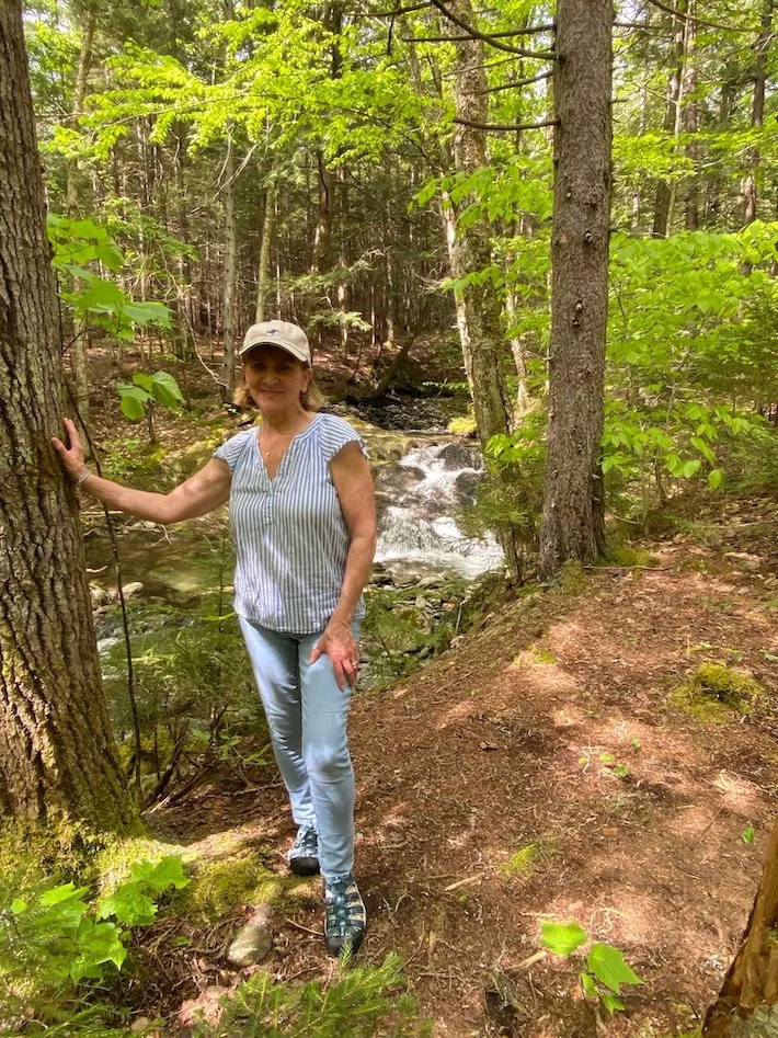 A woman standing on a dirt trail in a lush forest near a small creek, wearing a beige cap, striped blouse, and light-colored pants.