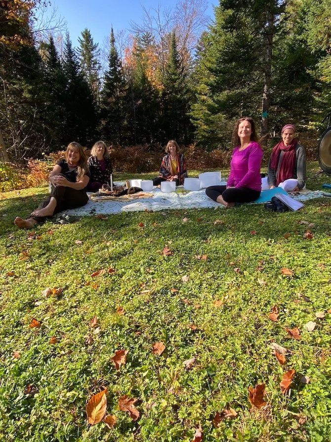 Five women sitting on a blanket outdoors in a wooded area with fall foliage, enjoying a picnic or gathering.