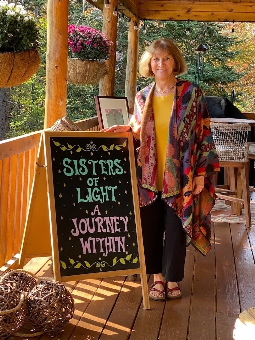 A woman standing on a wooden deck next to a sign that reads 'Sisters of Light, A Journey Within.' She is smiling, wearing a colorful patterned shawl, a yellow top, and black pants. There are hanging flower baskets and trees in the background, and the scene is lit with string lights.