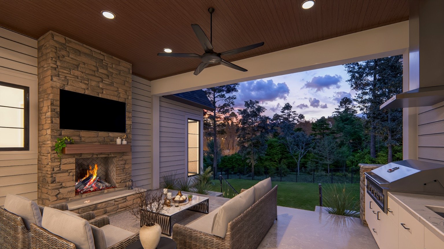 Covered outdoor patio with a stone fireplace, wall-mounted TV, ceiling fan, wicker furniture with cushions, a glass-top coffee table, and a grill, overlooking a backyard with trees at sunset.