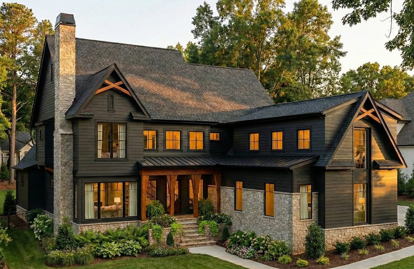 A custom luxury home by House of Severn featuring a modern farmhouse exterior with charcoal siding, natural stone masonry, and exposed timber trusses. The front elevation blends warm interior lighting, steep gabled rooflines, and layered landscaping 