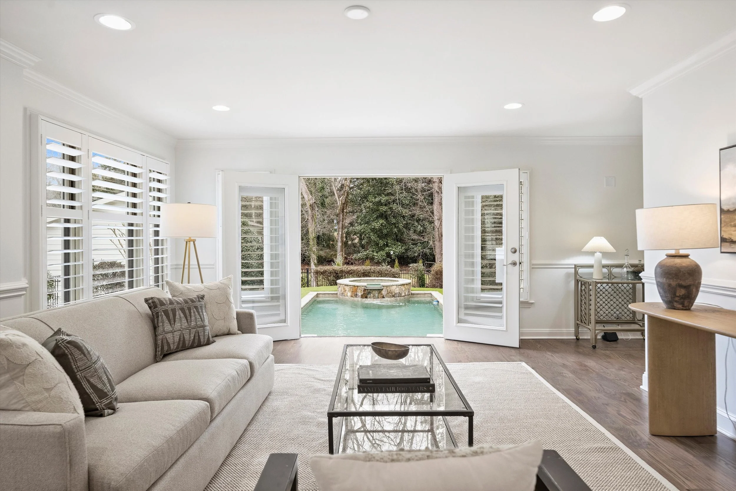 Light-filled family room with French doors opening to saltwater pool and spa, hardwood floors, neutral seating, and plantation shutters