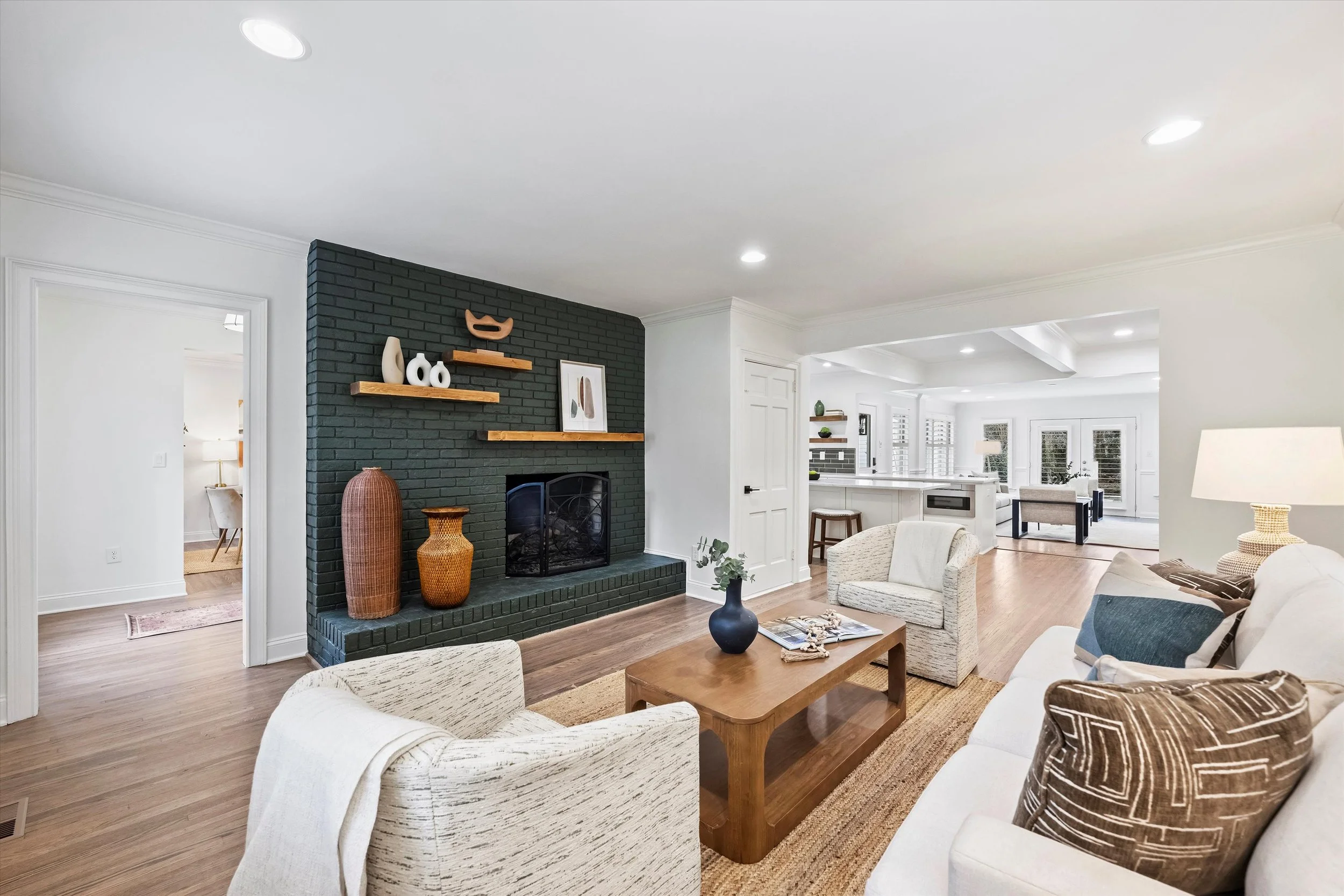 Bright open-concept living room with painted brick fireplace, floating wood shelves, neutral seating, hardwood floors, and view into modern white kitchen with island and tray ceiling