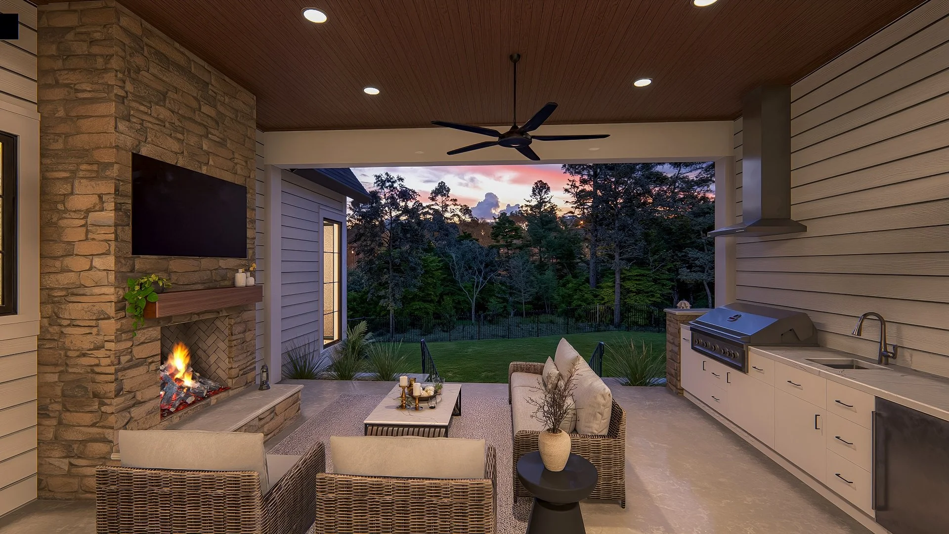 Covered outdoor patio with a fire pit, wicker seating, a large flat-screen TV mounted on a stone fireplace, a kitchen area with a grill, sink, and cabinets, and a view of a backyard with trees and a sunset sky.