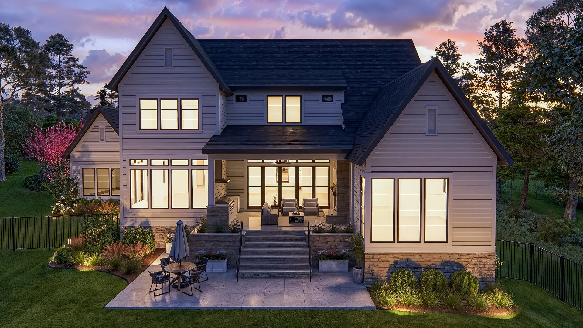 Beautiful modern house during dusk with large illuminated windows and a landscaped backyard patio featuring outdoor seating and a round table with an umbrella.