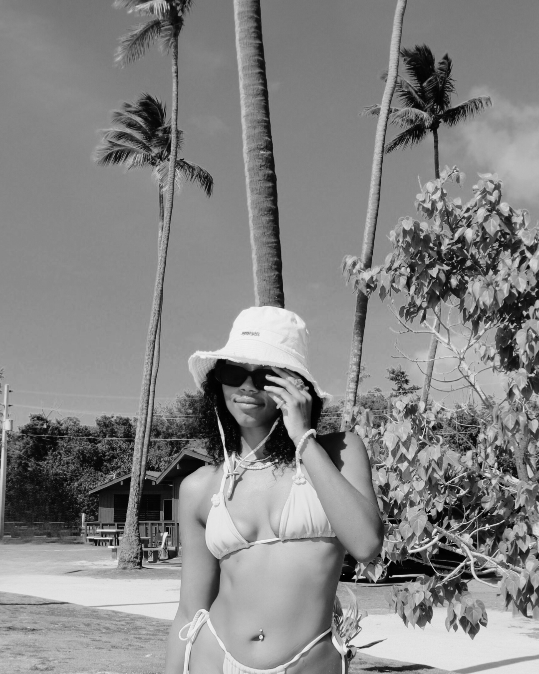 A woman wearing a bikini, sunhat, and sunglasses standing outdoors near palm trees and a small building.