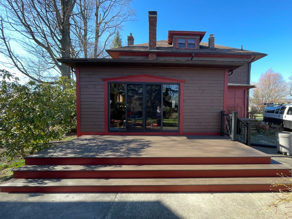 Back of a house with a wooden deck, sliding glass doors, and a small ramp with a black railing leading up to the deck.