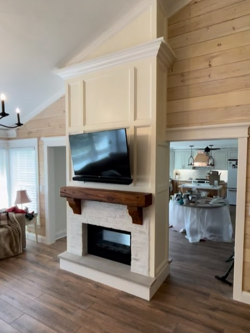 Living room with a mounted flat-screen TV above a white brick fireplace with a wooden mantle, beige wall paneling, a window with blinds, and a dining area visible through a doorway.