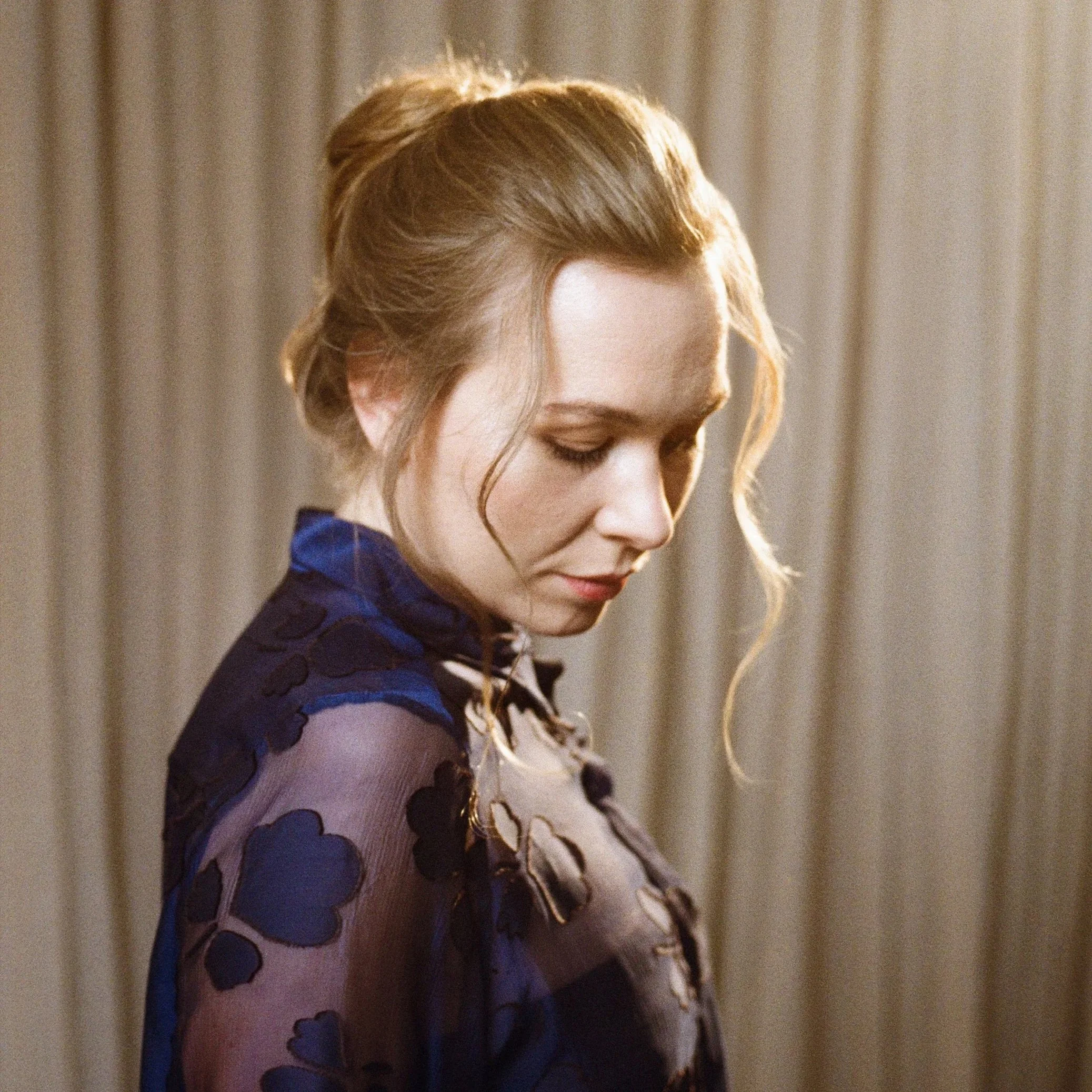 Elsa Dreisig with blonde hair styled in an updo, wearing a blue see-through dress with floral patterns, looking down with a soft expression, standing in front of beige curtains.