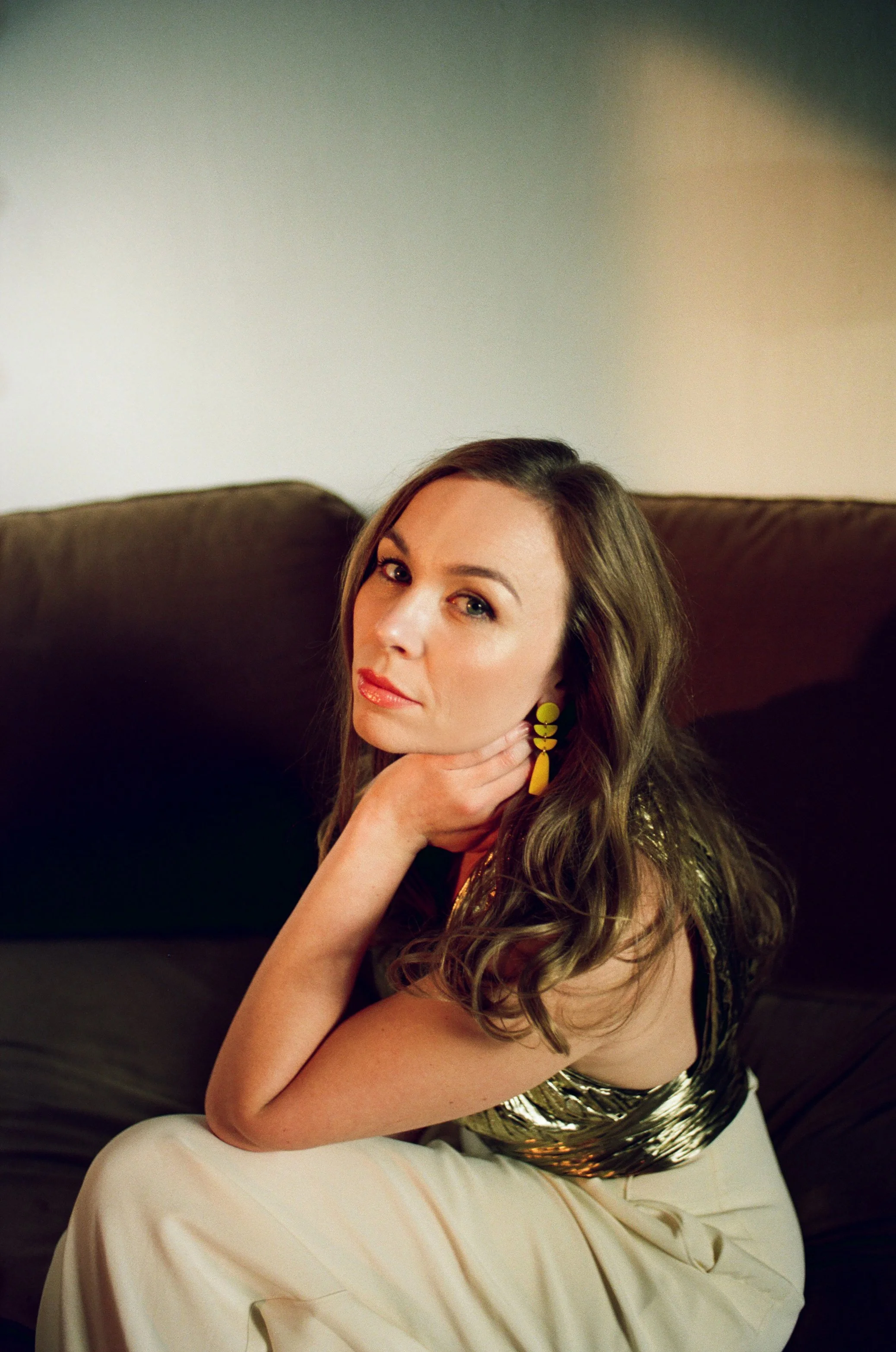 Elsa Dreisig singer with long wavy brown hair wearing a gold and black sleeveless top and cream-colored pants, sitting on a dark-colored sofa, resting her chin on her hand, looking at the camera.